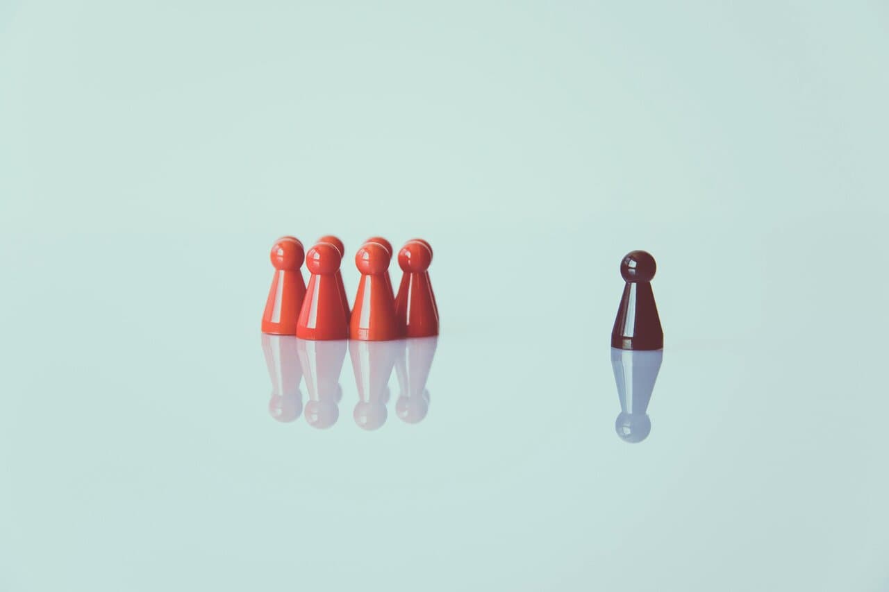 A single dark game piece stands separated from a clustered group of red game pieces on a reflective surface, symbolising isolation or individuality.