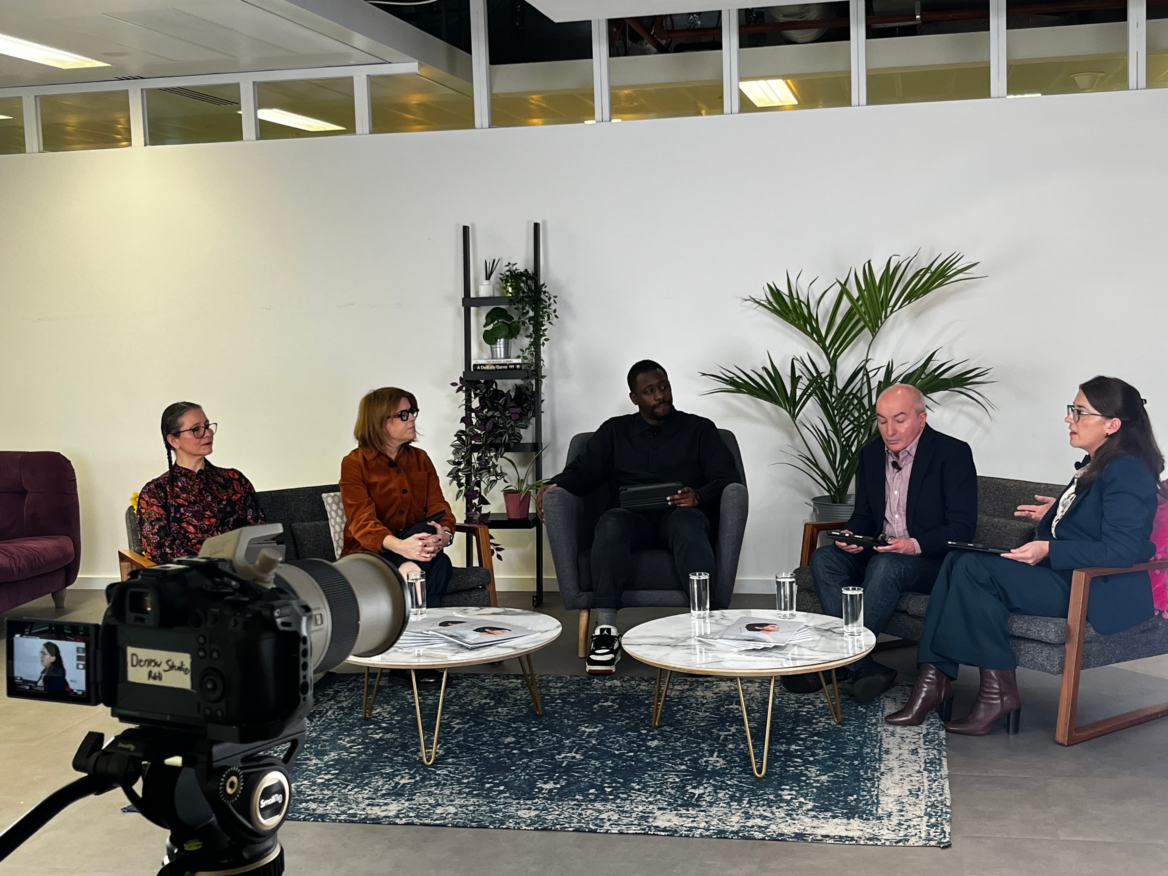 A panel of five people seated in a studio-style setting, engaged in conversation during a filmed discussion. A large softbox light is visible in the foreground on the left, and a camera setup appears on the right. The panelists sit on armchairs with small round tables in front of them, with plants and shelves in the background, creating a modern and professional atmosphere.